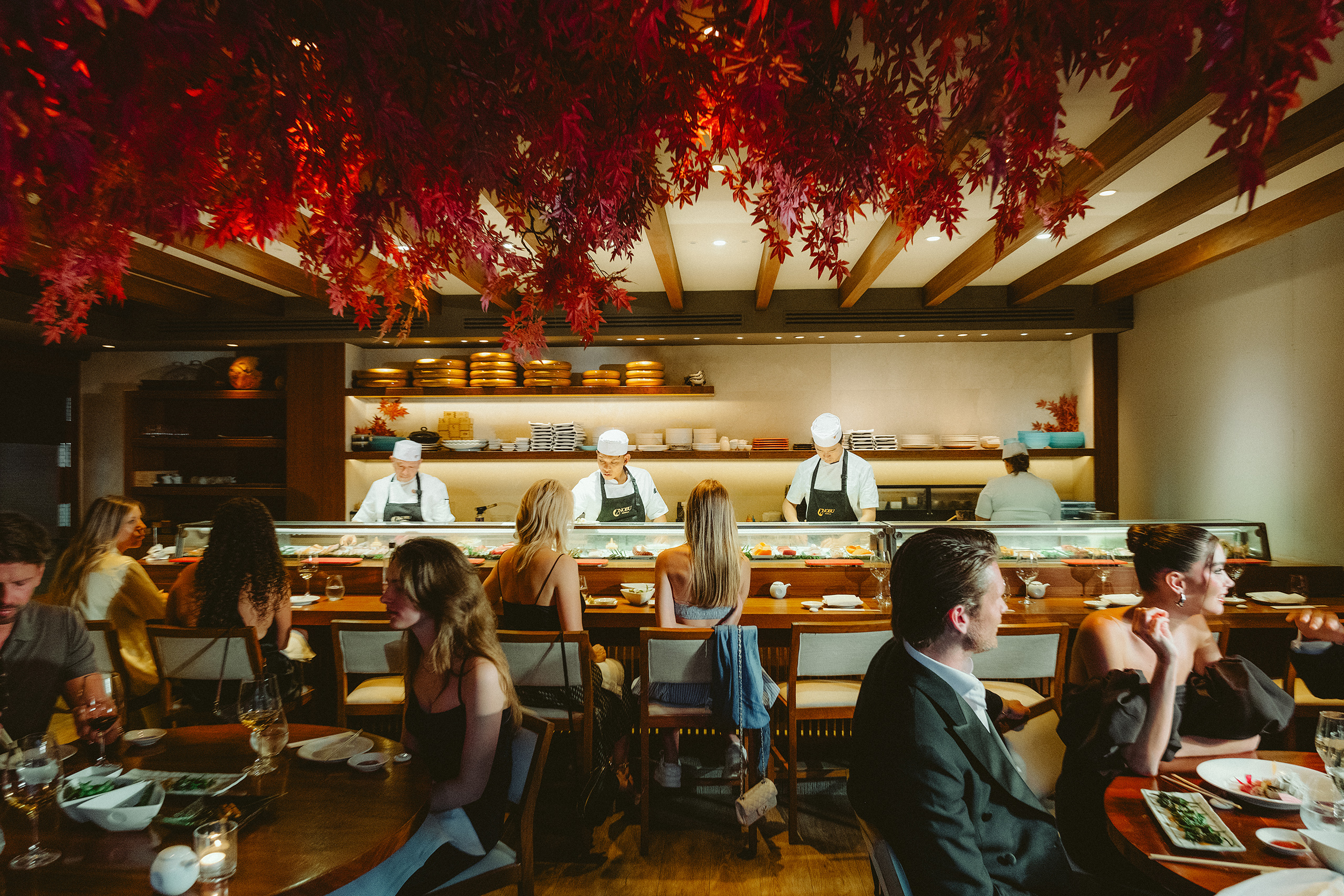 A lively restaurant filled with diners at tables, featuring beautiful red leaves draping from the ceiling above