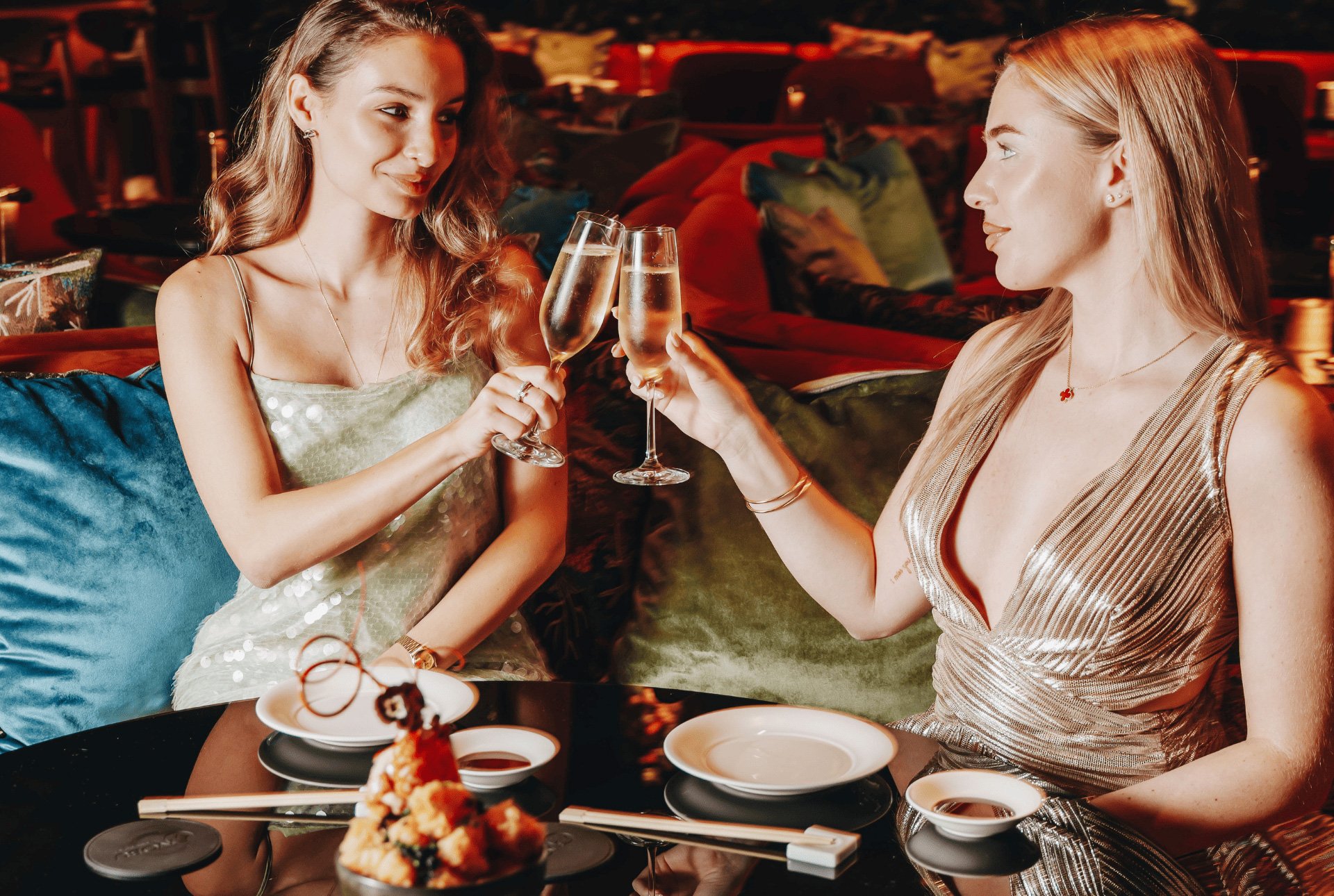 Two women in lovely dresses toast with champagne at a restaurant, sharing laughter and a delightful celebration.