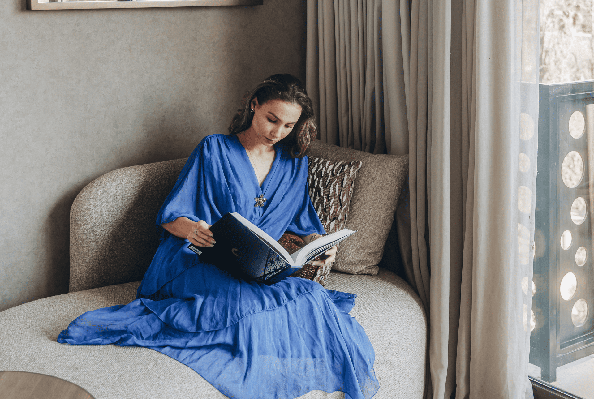 A woman wearing a blue dress relaxes on a couch, deeply focused on her book.