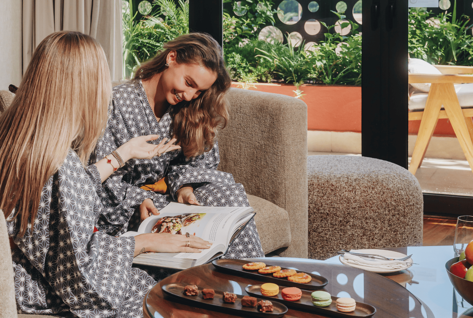 Two friends in pajamas relax on a couch, engrossed in a book, sharing smiles and a cozy atmosphere.