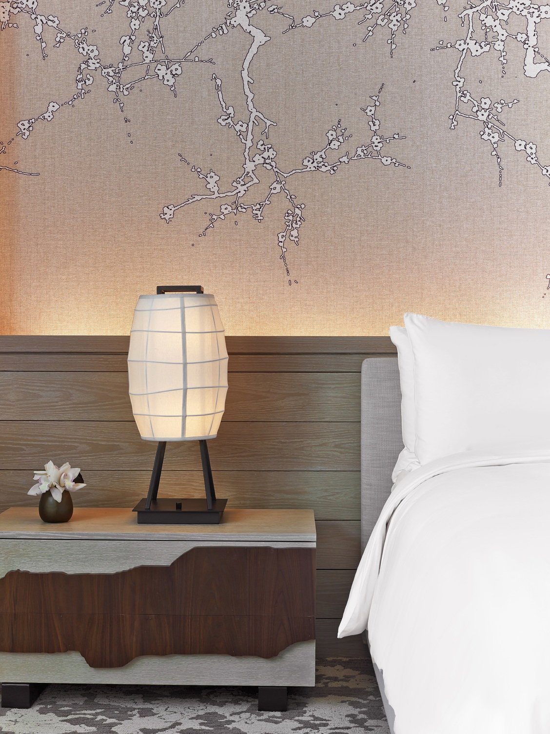 Wooden toned bed table, lamp, flower, and bed behind the Japanese styled wallpaper