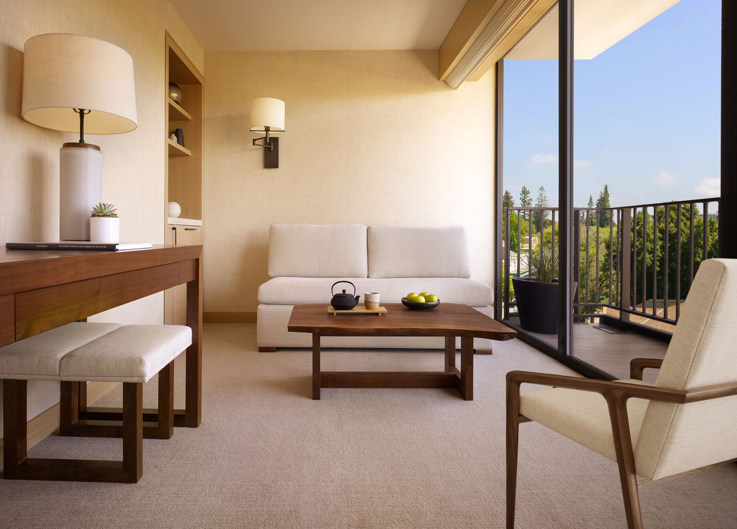 The junior suite shows a comfortable couch with table with a cup of tea, bowl of fruit, a desk area and a seat in the right corner. The room overlooks a balcony on a sunny day.